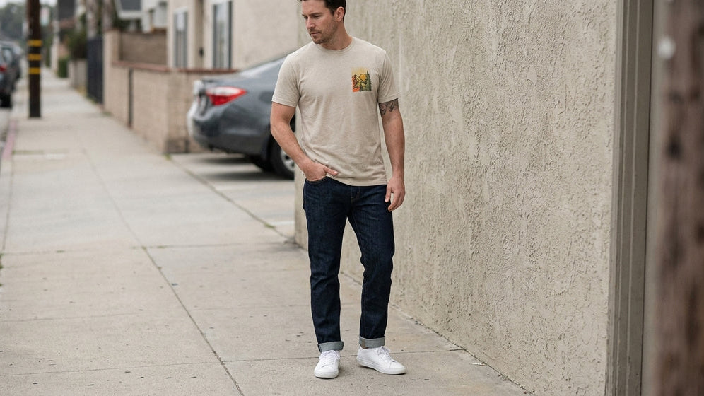 Man wearing a natural graphic tee with dark denim and white leather sneakers on a city sidewalk