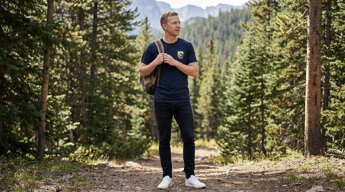 Man in the Campgrounds graphic tee in midnight navy adjusting his backpack strap at a forest trailhead